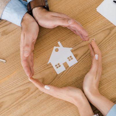 Couple making circle with hands on wooden table with paper house inside, house insurance